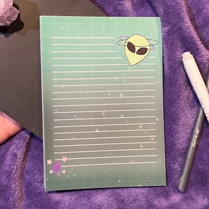 May include: A green notepad with a grid pattern and white lines. The notepad has a cartoon alien with a green head and two black eyes in the top right corner. The alien is surrounded by a circle of pink and yellow stars. The notepad has a pink mushroom with a white stem in the bottom left corner.