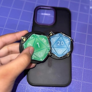 May include: Two phone grips with a dice design. One is green with the number 1, and the other is blue with the number 20. Both have a black base and are attached to a black phone case with a purple border. The background is a purple surface.