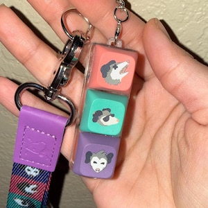 May include: A keychain featuring three colorful, square charms with opossum illustrations. The charms are pink, teal, and purple, and are attached to a silver clasp and a purple lanyard with a repeating pattern.