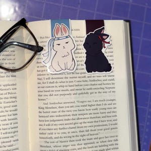 May include: Two decorative bunny bookmarks. One is white with a blue headband, and the other is black with a red bow. They are placed on the pages of an open book.