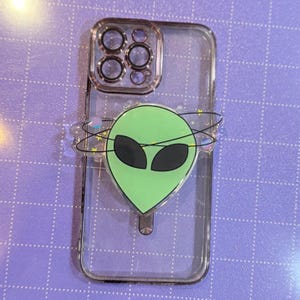 May include: A clear phone case with a green alien sticker on the back. The alien has black eyes and is surrounded by a glittery ring.