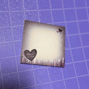 May include: A square sticker with a gradient background, transitioning from a light center to a darker border. The sticker features a heart with the text "Morally Grey" and a cityscape silhouette at the bottom.