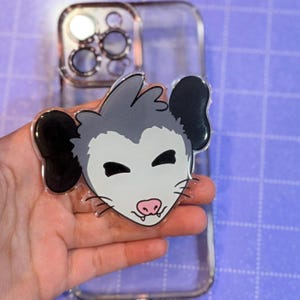 May include: A clear phone case with a cartoon opossum design. The opossum has a gray head, black ears, and a white face with a pink nose. The case is transparent, allowing the phone's color to show through.