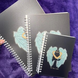 K&ouml;nnte beinhalten: Drei schwarze Spiralbl&ouml;cke mit verschiedenen Designs. Das obere Notizbuch hat ein blaues und wei&szlig;es Engelsfl&uuml;gel-Design. Das mittlere Notizbuch hat ein blaues und wei&szlig;es Engelsfl&uuml;gel-Design. Das untere Notizbuch hat eine schwarze Katze mit blauen und wei&szlig;en Engelsfl&uuml;geln und den Text "Fill the void".