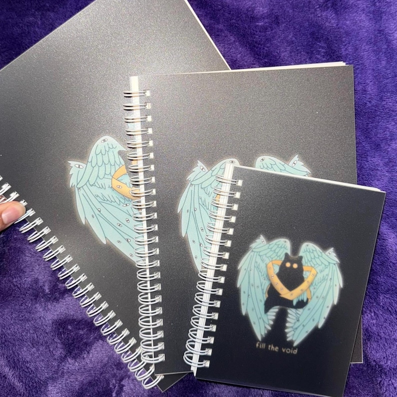 K&ouml;nnte beinhalten: Drei schwarze Spiralbl&ouml;cke mit verschiedenen Designs. Das obere Notizbuch hat ein blaues und wei&szlig;es Engelsfl&uuml;gel-Design. Das mittlere Notizbuch hat ein blaues und wei&szlig;es Engelsfl&uuml;gel-Design. Das untere Notizbuch hat eine schwarze Katze mit blauen und wei&szlig;en Engelsfl&uuml;geln und den Text "Fill the void".