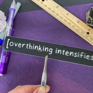 May include: A black sticker with white text that reads "[overthinking intensifies]" on a purple surface.