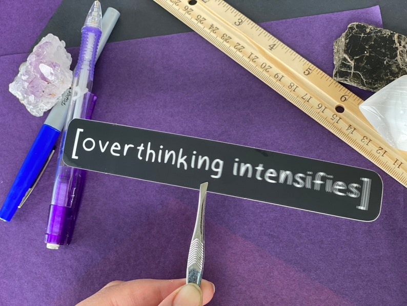 May include: A black sticker with white text that reads "[overthinking intensifies]" on a purple surface.