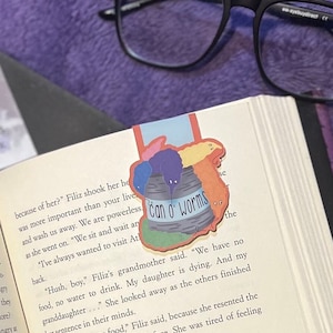 May include: A book with a bookmark featuring a cartoon illustration of a can of worms with the text "Can o' Worms". The bookmark is placed on a page with text about a grandmother and her granddaughter.