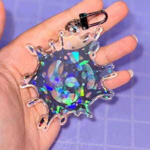 Puede incluir: Un llavero transparente e iridiscente con una forma de salpicadura única. El llavero presenta un interior oscuro y moteado con acentos holográficos coloridos. Tiene un cierre de metal negro.