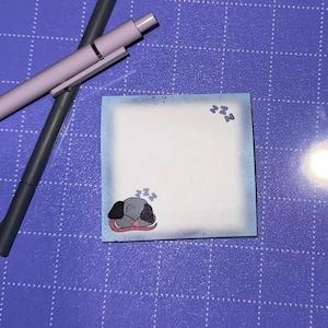 Könnte beinhalten: Quadratischer Haftzettel mit hellblauem Rand und einer Illustration eines schlafenden Hundes. Der Hund ist grau mit schwarzen Ohren und liegt auf einer rosa Oberfläche. Auf dem Zettel steht das Wort "zzz". Zwei Stifte befinden sich im Hintergrund.