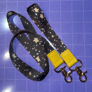 May include: A black lanyard with a repeating pattern of white and tan stars. The lanyard has yellow leather accents and silver-colored metal clasps. The lanyard is designed to be worn around the neck.