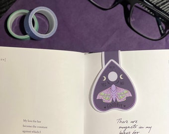 May include: A purple and green ouija board bookmark with a moth and the words "Hello", "Yes", "No", and "Goodbye". The bookmark is on a page of a book with the text "My love for her became the constant against which I measured truth." and "There are magnets in my bones for the ion in her blood."
