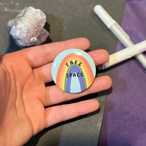 Puede incluir: Un botón de colores arcoíris con las palabras "SAFE SPACE" en texto negro.