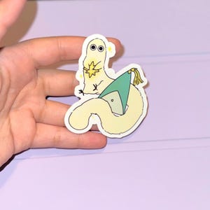 May include: A die-cut sticker featuring a whimsical cartoon character with a yellow body, holding a star. The character is paired with a worm-like creature wearing a green hat. The sticker has a white border and is set against a light purple background.