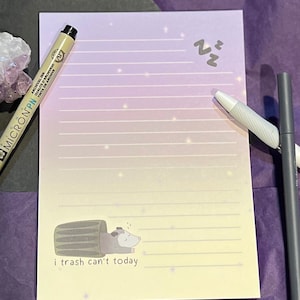 May include: A light pink and yellow lined notepad with a cute illustration of a possum sleeping in a bin. The text "i trash can't today" is printed below the illustration. The notepad has a white background with a subtle sparkle effect.