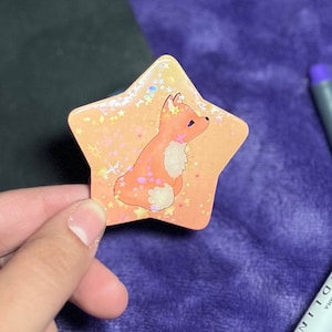 May include: A star-shaped sticker with a holographic finish features a cartoon fox with orange fur and a white chest. The fox is sitting on a peach-colored background with a scattering of gold stars.
