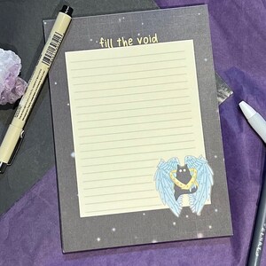 Puede incluir: Un bloc de notas blanco y negro con un gato negro con alas y un halo amarillo. El bloc de notas tiene un fondo negro con estrellas blancas y el texto "fill the void" en amarillo.