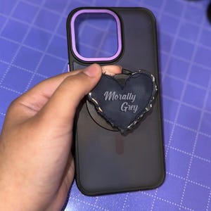 May include: A dark gray phone case with a purple camera ring and a heart-shaped phone grip. The phone grip is dark gray with the words "Morally Grey" in a light gray script.