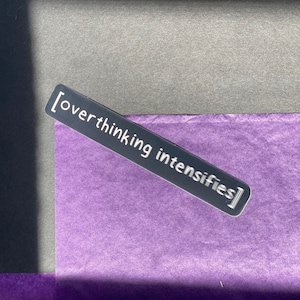 May include: A silver sticker with the text "[overthinking intensifies]" on a purple background.