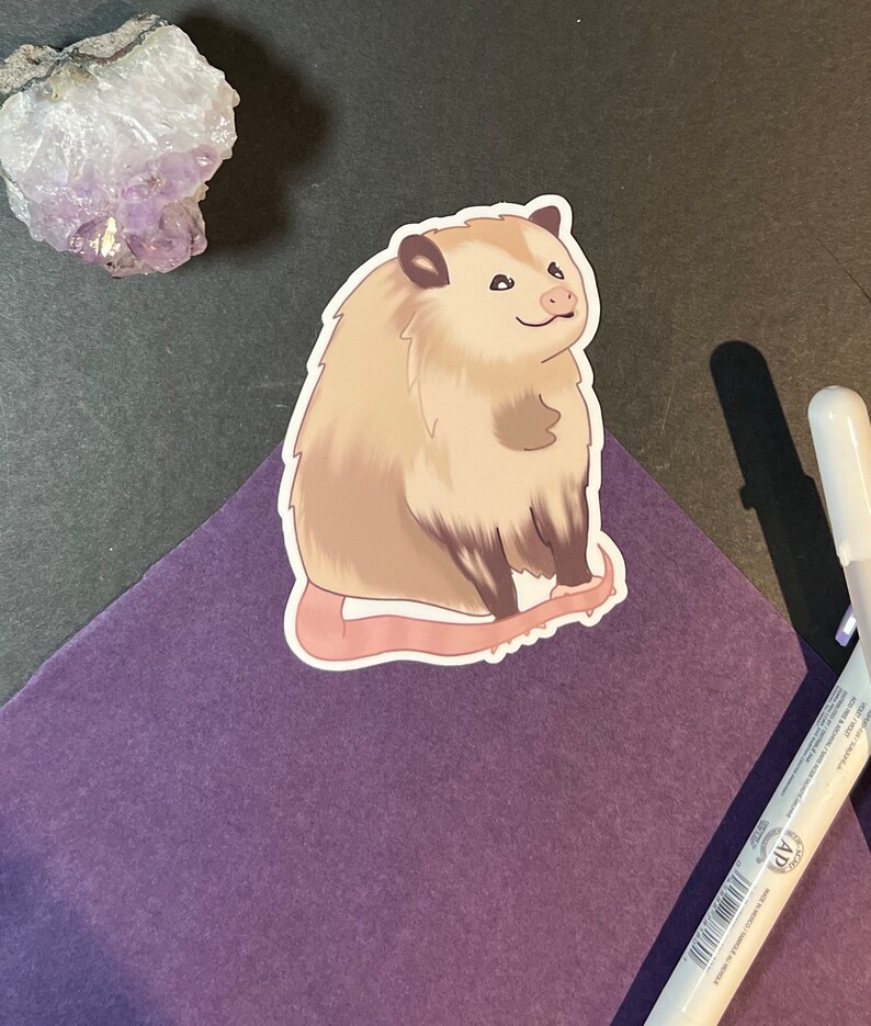 May include: A sticker of a cartoon opossum with a light brown body, a white face, and a pink nose. The opossum is sitting on its haunches with its tail curled around its body.