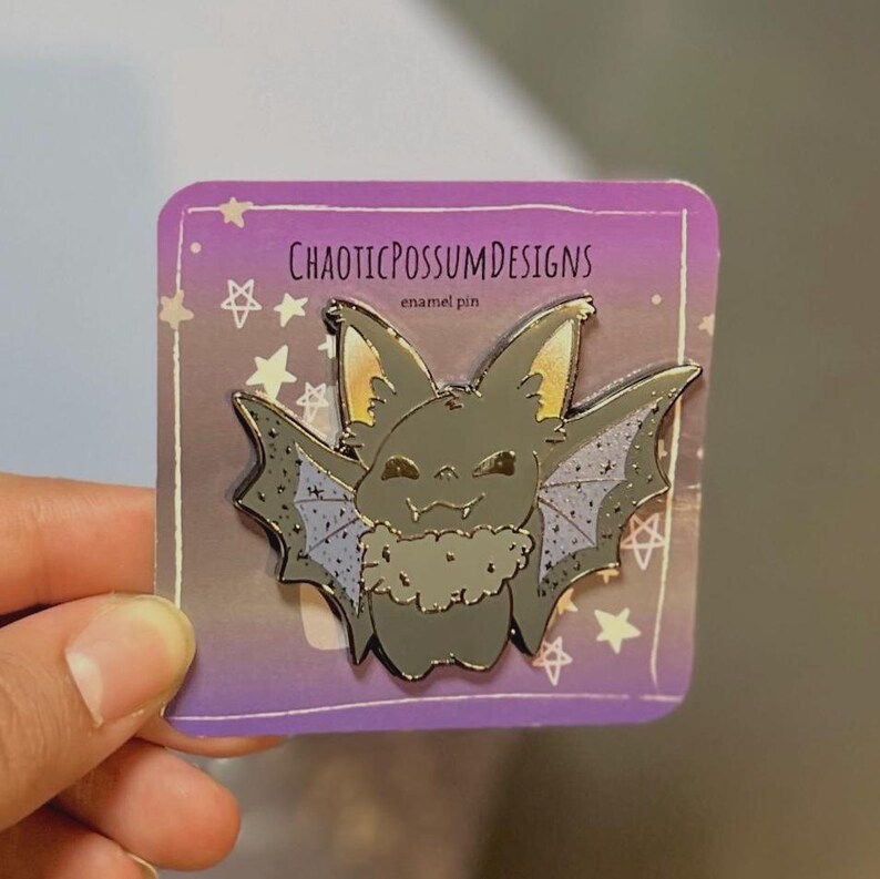 May include: A black enamel pin featuring a cartoon bat with a fluffy gray scarf. The pin is on a purple card with the text "CHAOTICPOSSUMDESIGNS enamel pin" and stars.