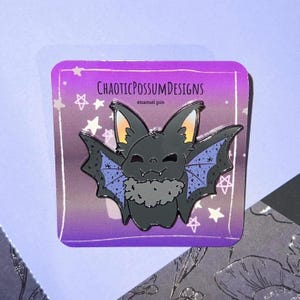 May include: A black enamel pin featuring a cartoon bat with purple wings and a fluffy gray collar. The pin is set against a purple and white gradient background with stars and pentagrams. The text "CHAOTICPOSSUMDESIGNS" and "enamel pin" are printed on the pin.