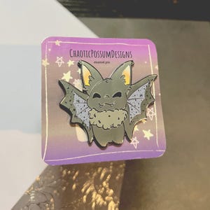 May include: A gray enamel pin featuring a cartoon bat with a fluffy white scarf. The bat has a purple and black patterned wing and a happy expression. The pin is on a purple background with stars and the text "CHAOTICPOSSUMDESIGNS enamel pin".