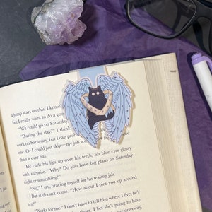 May include: A whimsical bookmark featuring a black cat with blue angel wings and a gold band. The bookmark is placed on an open book. A purple crystal and glasses are in the background. A pen is also visible.