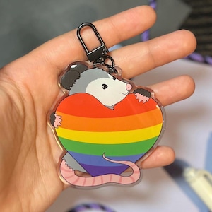 May include: A clear keychain with a cartoon possum holding a rainbow heart. The possum is gray with a pink nose and tail.