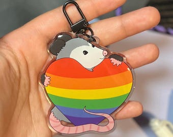 Puede incluir: Un llavero transparente con un opossum de dibujos animados sosteniendo un corazón arcoíris. El opossum es gris con la nariz y la cola rosas.