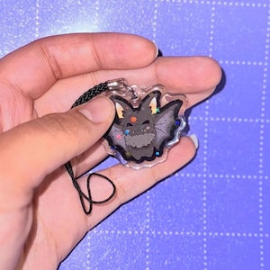Puede incluir: Un dije acrílico transparente con un murciélago de dibujos animados con una cara sonriente, cuerpo gris y alas negras. El dije tiene un cordón negro y está decorado con purpurina colorida en forma de estrella. El dije de murciélago es un accesorio lindo.