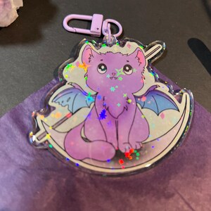 Pu&ograve; includere: Un gatto viola con le ali e una luna crescente dietro di lui. Il gatto ha una finitura scintillante e iridescente ed &egrave; seduto su un portachiavi in acrilico trasparente.