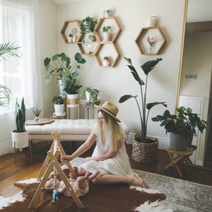 May include: A woman in a white dress and a straw hat plays with a baby on a brown and white cowhide rug. The room is decorated with plants and wooden hexagonal shelves.