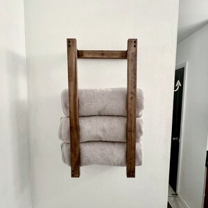 Puede incluir: Un toallero de madera con tres toallas dobladas y apiladas sobre &eacute;l. El toallero est&aacute; montado en una pared blanca.