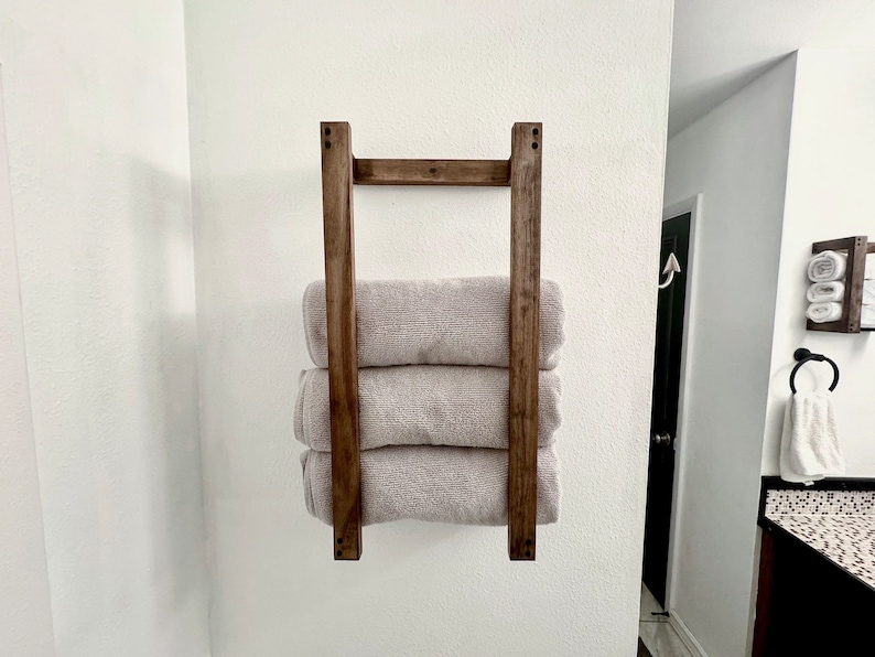 Puede incluir: Un toallero de madera con tres toallas dobladas y apiladas sobre &eacute;l. El toallero est&aacute; montado en una pared blanca.