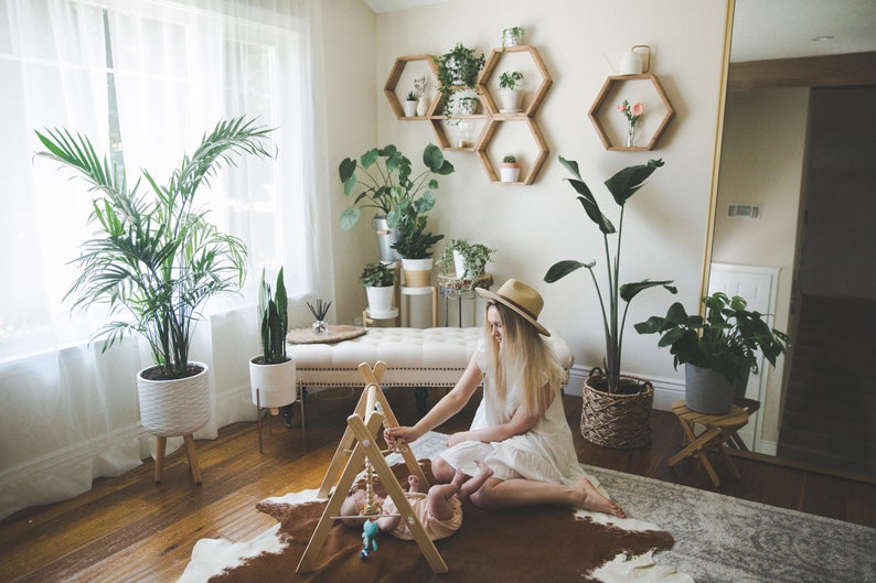 May include: A woman in a white dress and a straw hat plays with a baby on a brown and white cowhide rug. The room is decorated with plants and wooden hexagonal shelves.