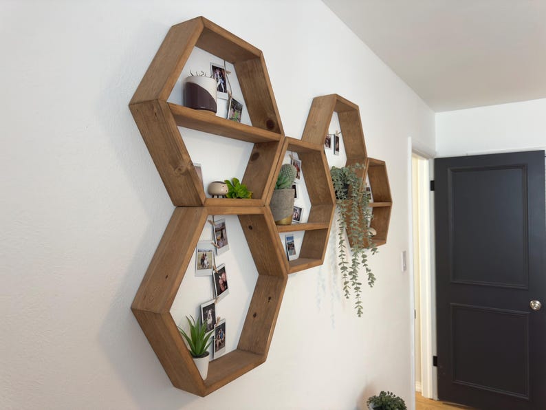 May include: A set of six hexagonal wooden shelves arranged in a honeycomb pattern on a white wall. The shelves are filled with small plants, photos, and other decorative items.