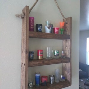 May include: A rustic wooden shelf with three shelves, hanging from the ceiling with rope. The shelves are filled with shot glasses of various colors and designs.
