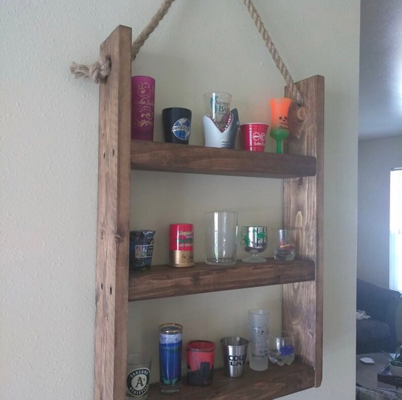 May include: A rustic wooden shelf with three shelves, hanging from the ceiling with rope. The shelves are filled with shot glasses of various colors and designs.