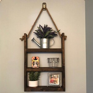 May include: A three-tiered wooden shelf with rope hanging from the top. The shelf is decorated with a small watering can, a wire basket, a picture frame, and a potted plant.