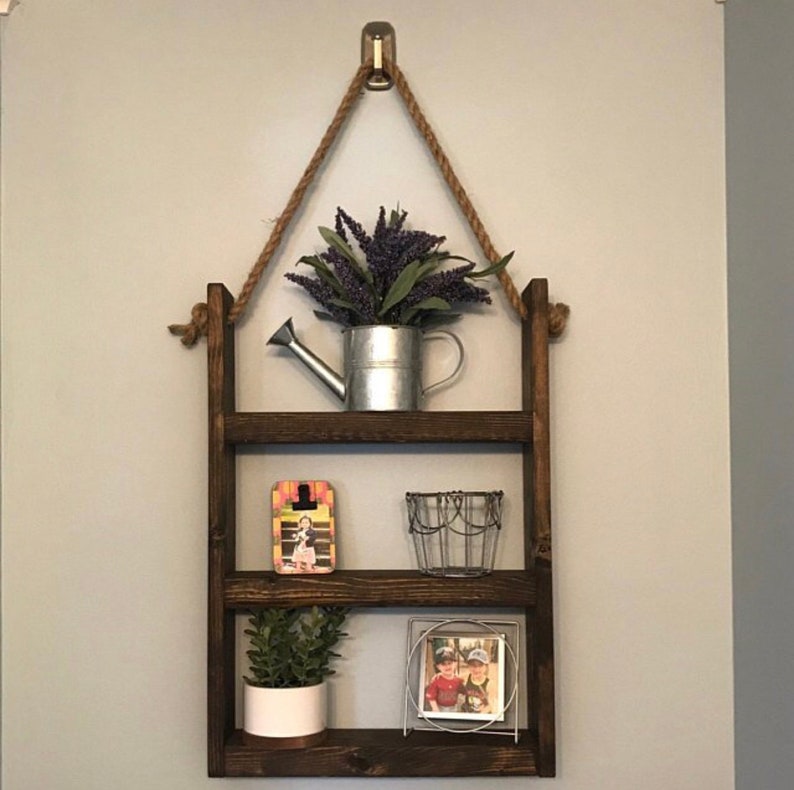 May include: A three-tiered wooden shelf with rope hanging from the top. The shelf is decorated with a small watering can, a wire basket, a picture frame, and a potted plant.