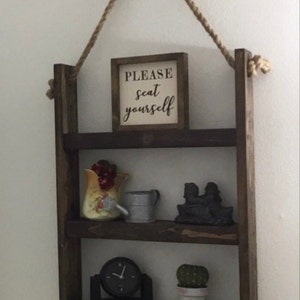 May include: A rustic wooden shelf with three tiers, hanging from a rope. The shelf is decorated with a sign that says "PLEASE seat yourself", a small watering can, a ceramic pitcher with fruit, a small cactus in a pot, and a clock.