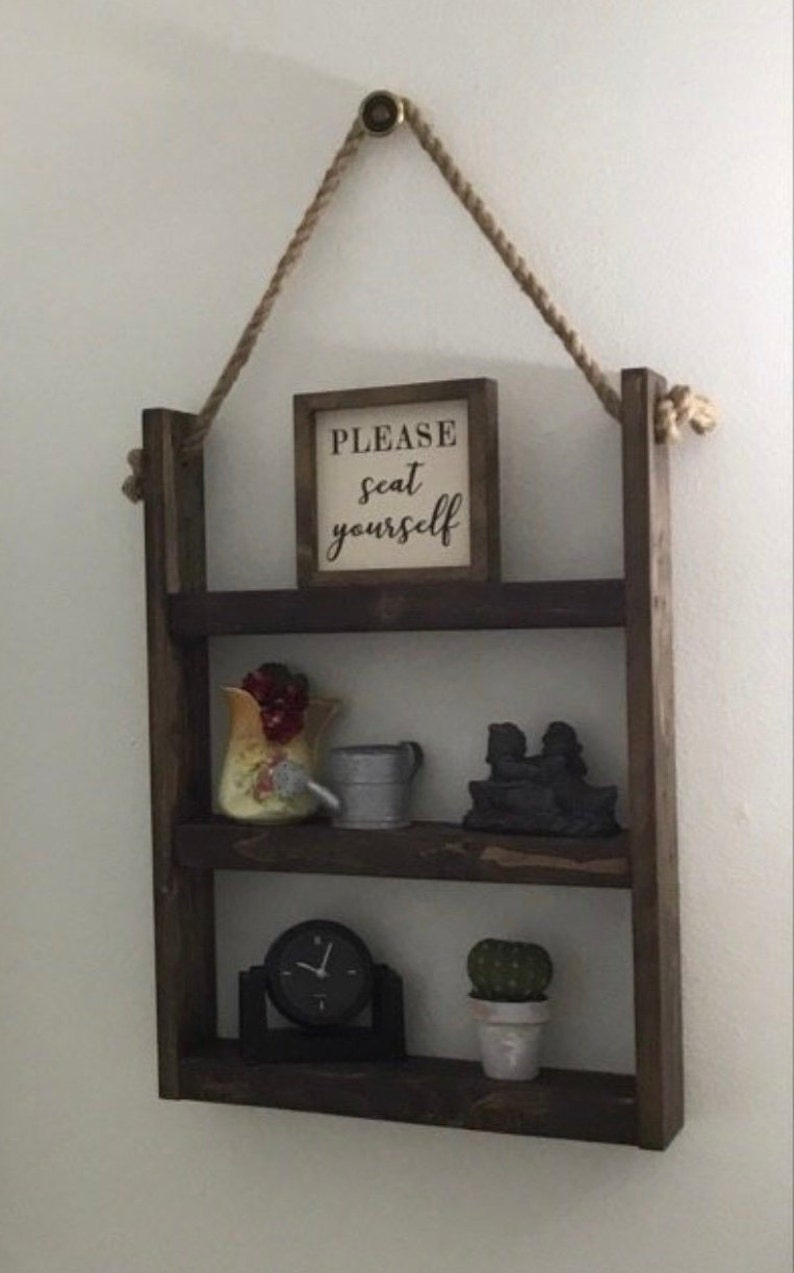 May include: A rustic wooden shelf with three tiers, hanging from a rope. The shelf is decorated with a sign that says "PLEASE seat yourself", a small watering can, a ceramic pitcher with fruit, a small cactus in a pot, and a clock.