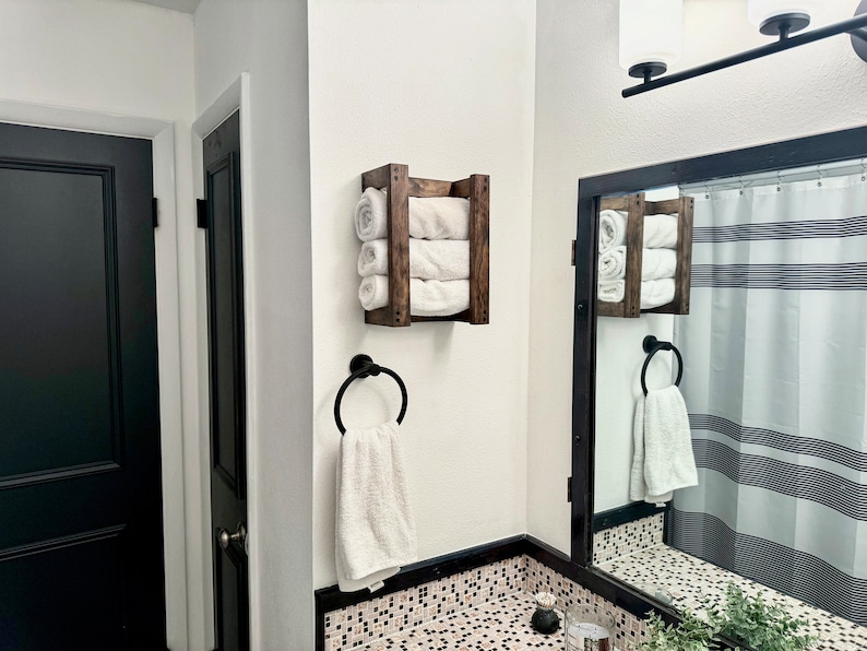 Puede incluir: Un toallero de madera con tres toallas blancas apiladas en &eacute;l. El toallero est&aacute; montado en una pared blanca en un ba&ntilde;o. Hay un anillo de toalla negro con una toalla blanca colgando.