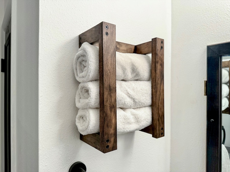 Puede incluir: Un toallero de madera con cuatro toallas blancas apiladas en su interior. El toallero est&aacute; montado en una pared blanca y tiene una mancha marr&oacute;n oscura.