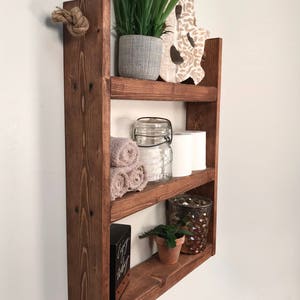 May include: A rustic wooden shelf with three tiers, hanging from a rope. The shelf is decorated with plants, towels, a glass jar, and a decorative candle holder. The shelf is stained a dark brown color.