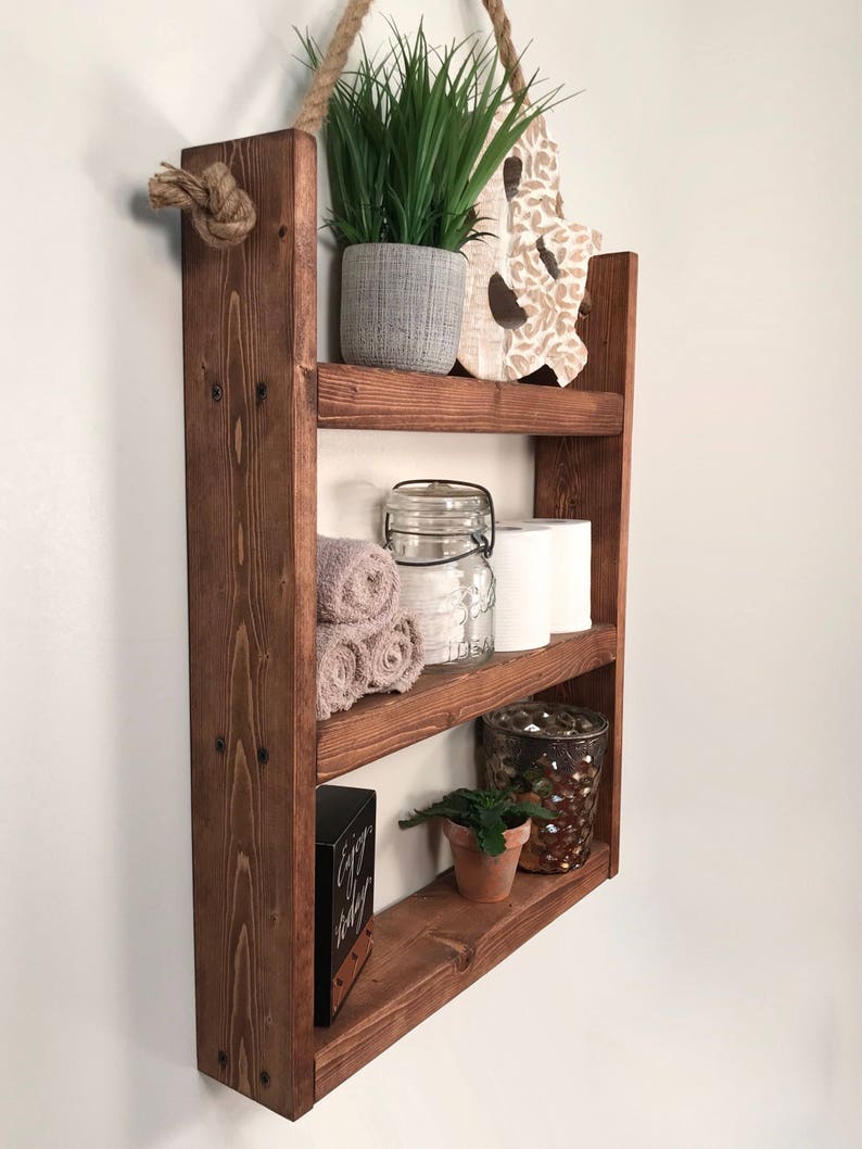 May include: A rustic wooden shelf with three tiers, hanging from a rope. The shelf is decorated with plants, towels, a glass jar, and a decorative candle holder. The shelf is stained a dark brown color.