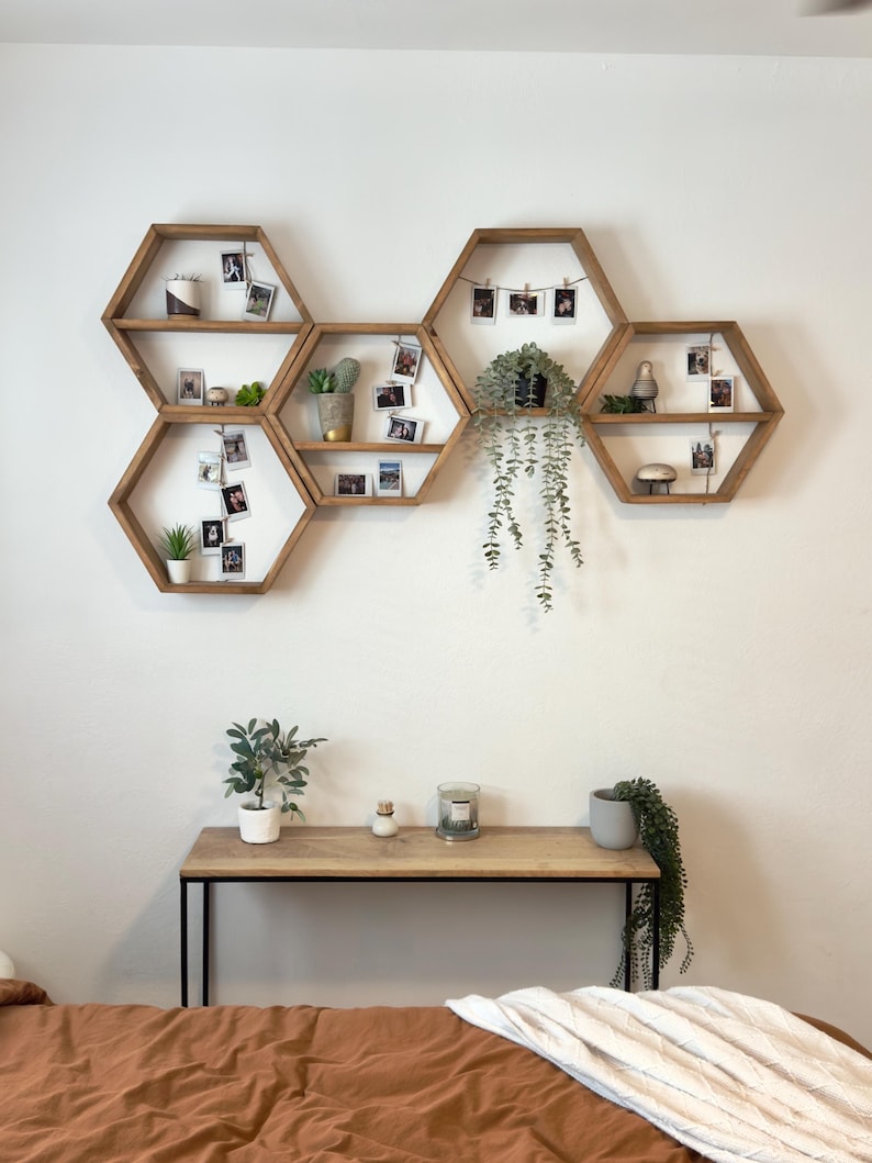 May include: A set of six hexagonal wooden shelves arranged in a honeycomb pattern on a white wall. The shelves are decorated with potted plants, small framed photos, and a string of lights. The shelves are mounted on a white wall. Below the shelves is a wooden table with black metal legs. The table is decorated with a potted plant, a candle, and a small white object. The table is in front of a brown bed with a white blanket.