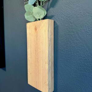 Op de afbeelding: Een houten wandplanter met een lichtbruine afwerking. De planter is gemonteerd op een donkerblauwe muur en heeft een takje groene eucalyptusbladeren erin.