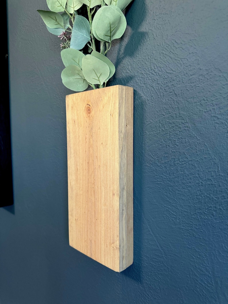 Op de afbeelding: Een houten wandplanter met een lichtbruine afwerking. De planter is gemonteerd op een donkerblauwe muur en heeft een takje groene eucalyptusbladeren erin.